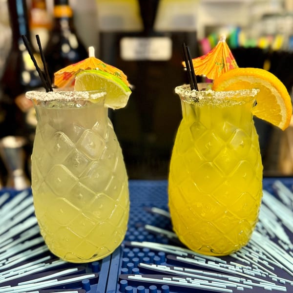 Two pineapple-shaped margaritas, one garnished with lime, the other with orange, on a blue bar mat.
