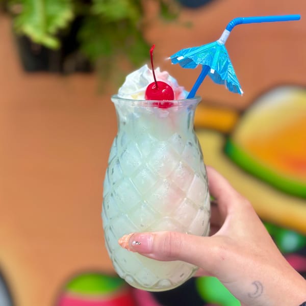 Hand holding a pineapple-shaped glass with a frozen drink, whipped cream, cherry, and blue umbrella.