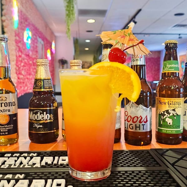 A colorful cocktail with an orange slice and cherry, surrounded by various beer bottles on a bar.