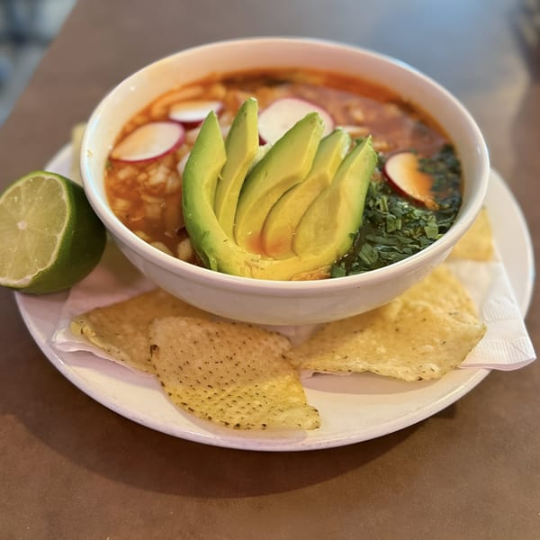 A bowl of Mexican pozole soup with avocado, radishes, cilantro, and a lime wedge, served with tortilla chips.