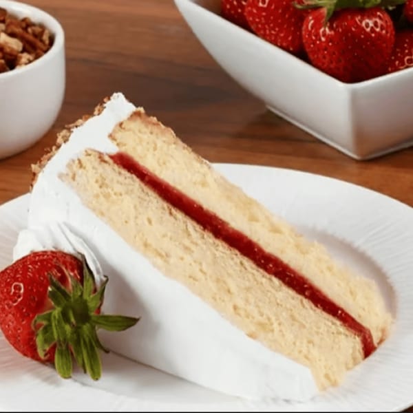 Slice of vanilla cake with strawberry filling and whipped cream, garnished with a fresh strawberry.