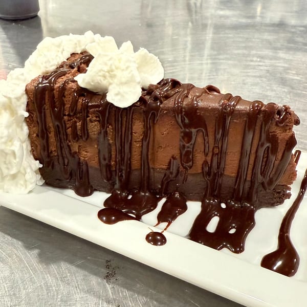 Rich chocolate cheesecake slice with chocolate drizzle and whipped cream on a white plate.