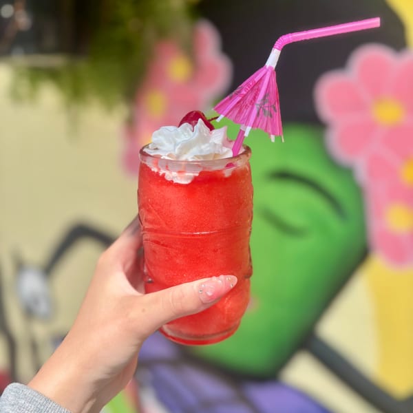 Hand holding a vibrant red frozen drink with whipped cream, a cherry, and a pink umbrella straw.